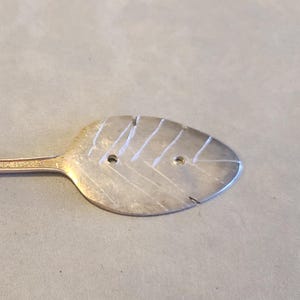 May include: A silver-toned metal wall hook fashioned from a repurposed spoon. The handle is curved into a hook shape with an ornate floral pattern, while the bowl of the spoon is flattened and etched with a leaf-like design featuring two holes.