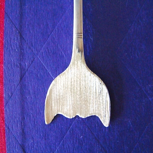 May include: A silver spoon with a mermaid tail design. The spoon is on a blue background.