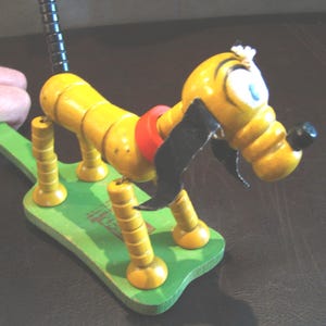 Puede incluir: Un juguete de madera vintage con forma de perro, pintado de amarillo con orejas negras y un collar rojo, montado sobre una base verde. El cuerpo del perro es articulado. El juguete está sostenido por una mano.