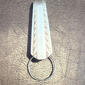 May include: A silver-toned metal keychain featuring a flat, tapered handle section from a piece of silverware. The handle has a decorative embossed border pattern along both sides and is attached to a circular metal split ring for keys.