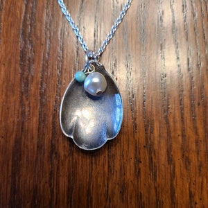 Puede incluir: Collar con colgante de cuchara de plata con detalles de perlas y cuentas turquesas. El cuenco de la cuchara tiene forma de concha de vieira. La cadena del collar es una cadena de trigo plateada.