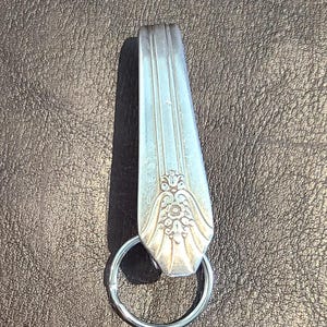 May include: A silver-toned metal keychain featuring the handle of a vintage spoon as the main component. The spoon handle has a tapered shape with three vertical lines and an embossed floral pattern at the base, attached to a circular metal ring.