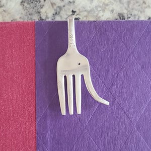 May include: A silver fork repurposed as a bookmark, with tines and a curved handle. The fork is placed on a purple book, with a pink section visible on the left. The fork's handle has decorative floral details.