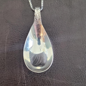 May include: A silver pendant necklace with a spoon-shaped charm. The charm has a reflective surface and is attached to a delicate chain. The pendant has an ornate design at the top.
