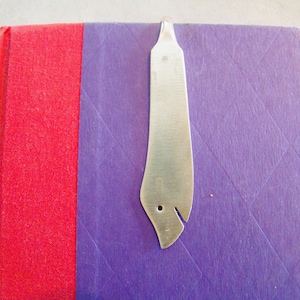May include: A silver metal bookmark shaped like a bird with a hole in the middle. The bookmark is on a purple and red book cover.