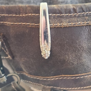 May include: Silverware spoon repurposed as a bag clip, attached to a brown leather bag. The spoon has a decorative floral design on the handle. The bag has visible stitching and a patterned fabric lining.