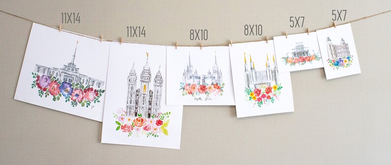 May include: A collection of watercolor prints featuring a white temple with colorful floral wreaths. The prints are displayed on a wall with clothespins and string. The prints are available in various sizes, including 5x7, 8x10, and 11x14 inches.