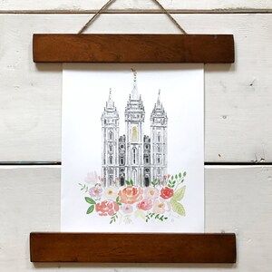 May include: A black and white illustration of a temple with a gold spire, surrounded by pink, white, and green watercolor flowers. The illustration is framed with a brown wooden frame.