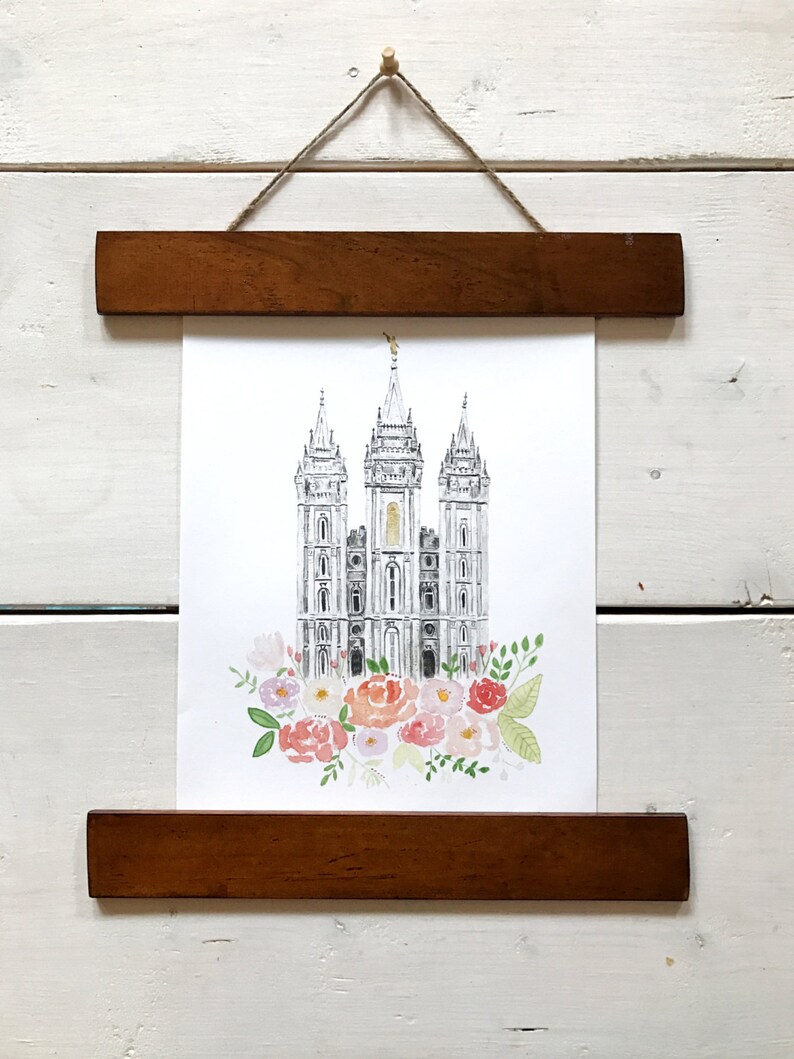 May include: A black and white illustration of a temple with a gold spire, surrounded by pink, white, and green watercolor flowers. The illustration is framed with a brown wooden frame.
