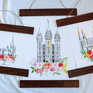 May include: Three watercolor prints of different temples with floral arrangements at the bottom. The prints are framed with dark wood and hung with twine.