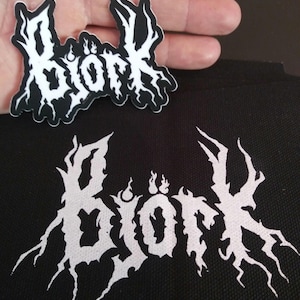 May include: A black and white patch with the word "Glor", stylized in a metal band logo font. The patch is on a black fabric background.