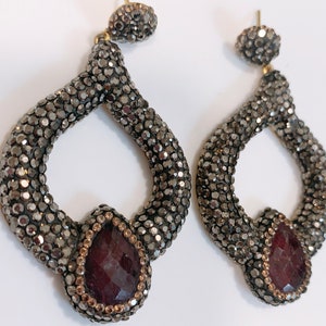 May include: A pair of dangle earrings with a teardrop-shaped red gemstone set in a silver-toned metal frame. The frame is covered in small, sparkling rhinestones.