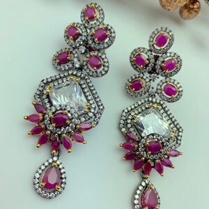 May include: A pair of ornate drop earrings featuring a large, clear, rectangular center stone surrounded by smaller white stones. The earrings have clusters of round, ruby-colored stones and a teardrop-shaped ruby at the bottom. The metal has a dark, gunmetal finish.