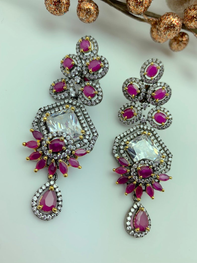 May include: A pair of ornate drop earrings featuring a large, clear, rectangular center stone surrounded by smaller white stones. The earrings have clusters of round, ruby-colored stones and a teardrop-shaped ruby at the bottom. The metal has a dark, gunmetal finish.