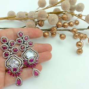 May include: A pair of silver-toned earrings with a geometric design. Each earring features a large, clear, square-cut stone surrounded by smaller white stones and ruby-colored accents. The earrings have a drop-style design with a teardrop-shaped ruby at the bottom.