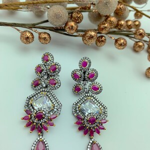 May include: A pair of ornate drop earrings featuring a geometric design. The earrings have a silver-tone metal setting with clear and ruby-colored stones. The earrings have a teardrop-shaped ruby at the bottom.
