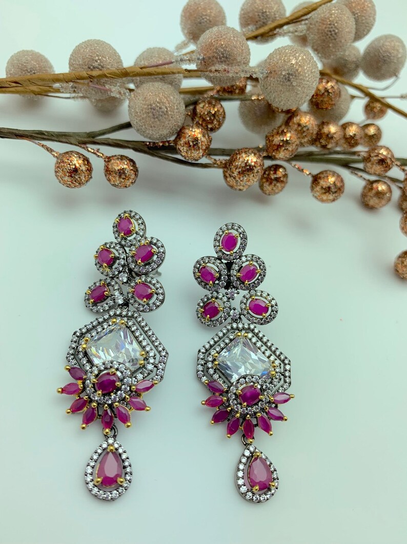 May include: A pair of ornate drop earrings featuring a geometric design. The earrings have a silver-tone metal setting with clear and ruby-colored stones. The earrings have a teardrop-shaped ruby at the bottom.