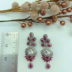 May include: A pair of ornate silver-toned earrings with ruby-red and clear crystal accents. The earrings feature a geometric design with a large, rectangular clear crystal at the center and a teardrop-shaped ruby at the bottom. The earrings measure approximately 4 inches in length.