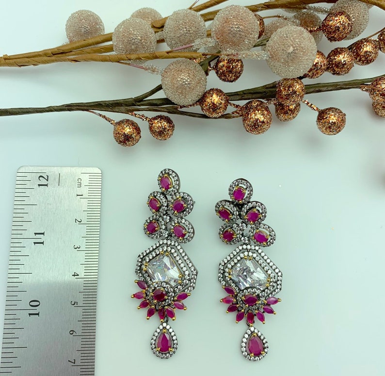 May include: A pair of ornate silver-toned earrings with ruby-red and clear crystal accents. The earrings feature a geometric design with a large, rectangular clear crystal at the center and a teardrop-shaped ruby at the bottom. The earrings measure approximately 4 inches in length.