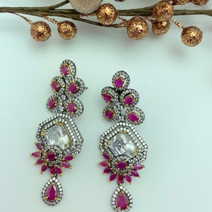 May include: A pair of ornate drop earrings. Each earring features a large, clear, rectangular stone surrounded by smaller white stones. Clusters of small, round, red stones and a teardrop-shaped red stone dangle below. The earrings have a dark metal setting.