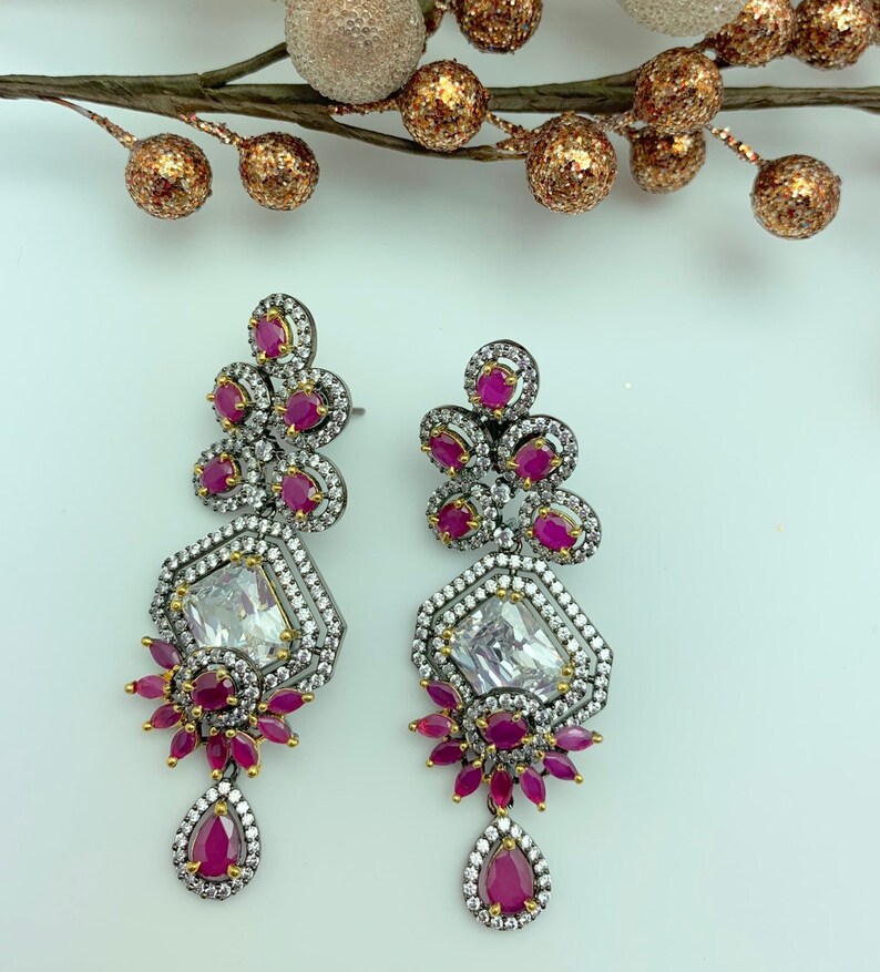 May include: A pair of ornate drop earrings. Each earring features a large, clear, rectangular stone surrounded by smaller white stones. Clusters of small, round, red stones and a teardrop-shaped red stone dangle below. The earrings have a dark metal setting.