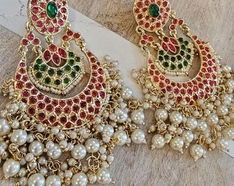 May include: A pair of ornate gold-toned earrings. Each earring features a circular top with a green center stone surrounded by red stones. A crescent-shaped design with red stones and a green center hangs below, with a cascade of white pearls.
