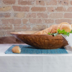 Puede incluir: Un cuenco de madera grande y ovalado con un patr&oacute;n de veta visible, lleno de varios trozos de pan y una guarnici&oacute;n de hojas verdes. Un trozo de pan descansa sobre un mantel individual rectangular de color verde azulado junto al cuenco.