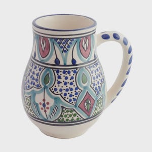 May include: A white ceramic mug with a handle, adorned with intricate blue, teal, pink, and green patterns. The handle features blue polka dots. The mug has a capacity of approximately 16 ounces.
