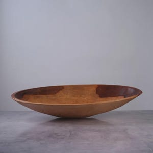 Puede incluir: Un cuenco de madera ancho y poco profundo con una silueta suave y curva. La superficie interior muestra un patr&oacute;n de veta de madera natural con una mancha marr&oacute;n oscura prominente en el lado derecho, que contrasta con los tonos bronceados claros.