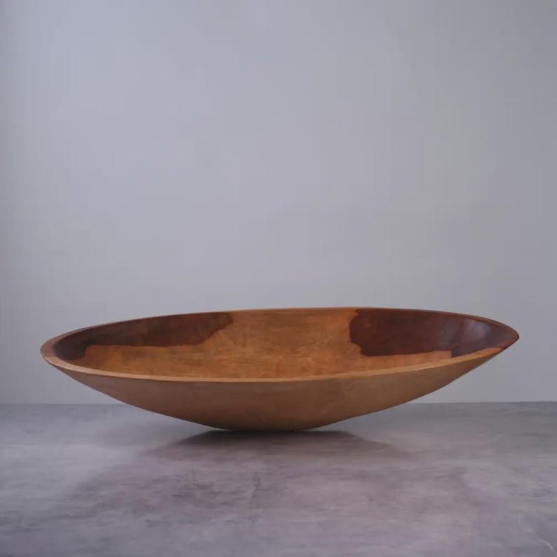 Puede incluir: Un cuenco de madera ancho y poco profundo con una silueta suave y curva. La superficie interior muestra un patr&oacute;n de veta de madera natural con una mancha marr&oacute;n oscura prominente en el lado derecho, que contrasta con los tonos bronceados claros.