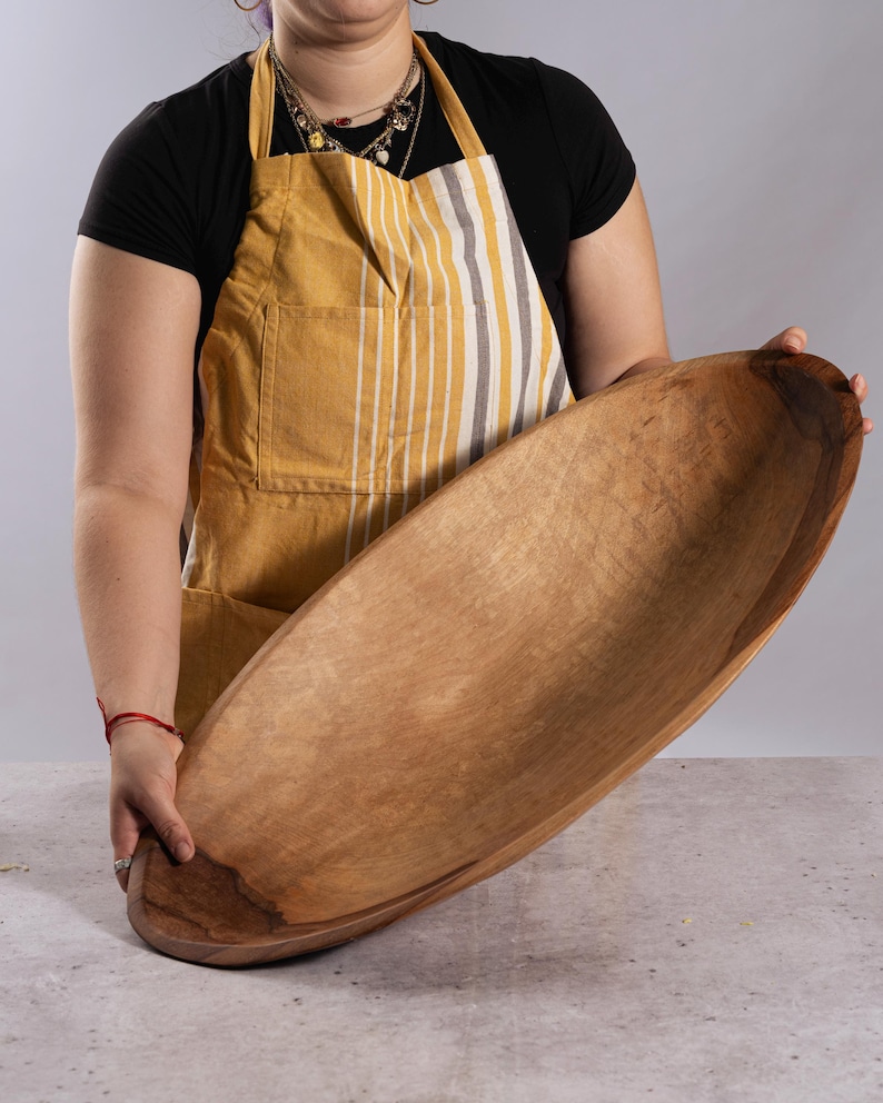 Puede incluir: Un cuenco de madera grande de forma ovalada con un interior liso de color marr&oacute;n claro y un patr&oacute;n de veta de madera natural m&aacute;s oscuro a lo largo del borde exterior. El cuenco presenta una cuenca profunda y alargada con un acabado pulido que resalta la textura org&aacute;nica.