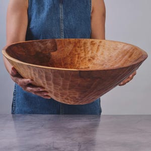 May include: A large, round wooden bowl with a natural light brown finish and a distinctively textured, hand-carved exterior surface. The bowl features visible wood grain patterns and a smooth, wide rim, showcasing a deep, open interior basin.