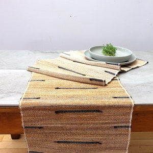 May include: A natural fiber table runner with a woven design featuring black stripes. The runner is folded in thirds and a light gray ceramic plate is on top.