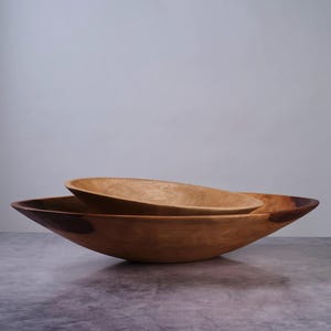 Puede incluir: Dos cuencos de madera encajados con un acabado liso y pulido y patrones de veta natural visibles. El cuenco m&aacute;s grande tiene un perfil ancho y poco profundo con acentos de color marr&oacute;n oscuro en el borde, mientras el peque&ntilde;o descansa dentro.