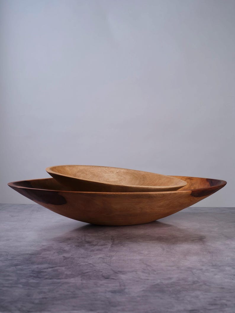 Puede incluir: Dos cuencos de madera encajados con un acabado liso y pulido y patrones de veta natural visibles. El cuenco m&aacute;s grande tiene un perfil ancho y poco profundo con acentos de color marr&oacute;n oscuro en el borde, mientras el peque&ntilde;o descansa dentro.
