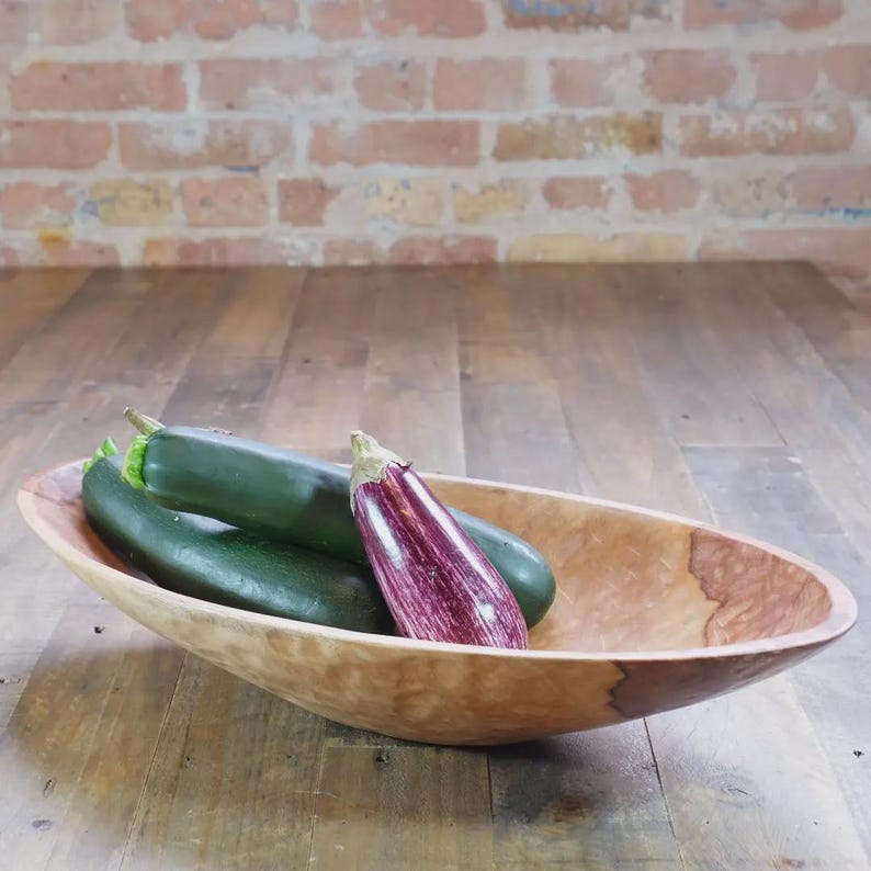 Puede incluir: Un cuenco de madera de forma ovalada con una veta de color marr&oacute;n claro natural y una mancha m&aacute;s oscura en el lateral. Dentro del cuenco hay dos calabacines de color verde oscuro y una peque&ntilde;a berenjena a rayas moradas y blancas.