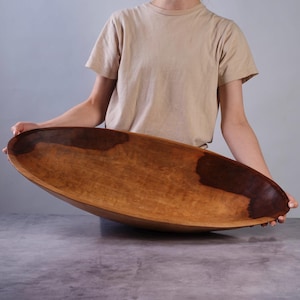 Puede incluir: Un cuenco de madera grande, alargado y de forma ovalada con un acabado liso y pulido. El cuenco presenta un patr&oacute;n de veta de madera natural con manchas de color marr&oacute;n oscuro que contrastan con la madera clara de tono miel c&aacute;lido.