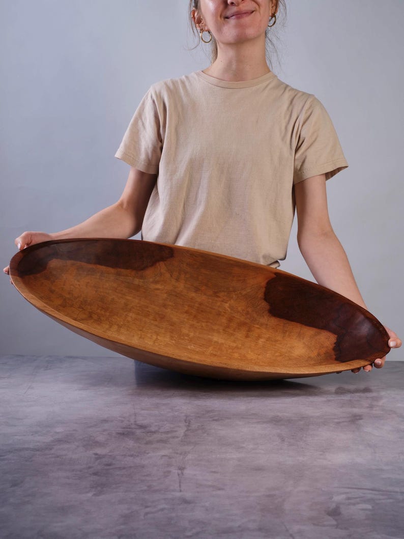 Puede incluir: Un cuenco de madera grande, alargado y de forma ovalada con un acabado liso y pulido. El cuenco presenta un patr&oacute;n de veta de madera natural con manchas de color marr&oacute;n oscuro que contrastan con la madera clara de tono miel c&aacute;lido.