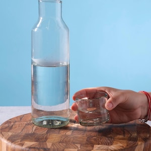 May include: A clear glass water bottle and a small glass tumbler, both partially filled with water. The bottle is tall and slender, while the tumbler is short and round. The items are on a wooden cutting board against a blue background.