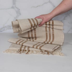May include: A rectangular woven table runner in a light beige color featuring vertical brown striped patterns. The fabric has a textured appearance and is finished with decorative fringe along the bottom edge, creating a simple and functional design.
