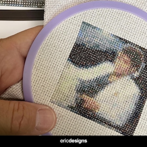 Pode incluir: Um close-up de um projeto de bordado de ponto cruz com uma pessoa usando uma camisa branca. O bordado está emoldurado em um aro roxo.