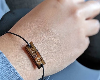 Pode incluir: Um bracelete de cordão preto com um amuleto retangular castanho que diz "Love" em itálico.