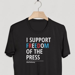 May include: Black t-shirt with white text that reads "I support freedom of the press" with the word "freedom" in red and blue. The hashtag "#SupportThePress" is printed below the text.