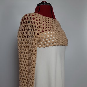 May include: A beige crochet shrug with a wide, open neckline and long sleeves. The shrug is made with a delicate open stitch pattern.