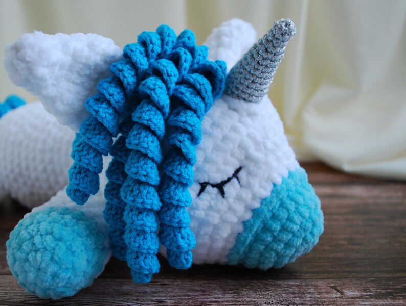 May include: A white crocheted unicorn with a blue yarn mane and tail, lying on a wooden surface. The unicorn has a silver glitter horn.