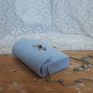 Puede incluir: Un bolso de peluche azul claro con una correa de cadena plateada y un cierre plateado.