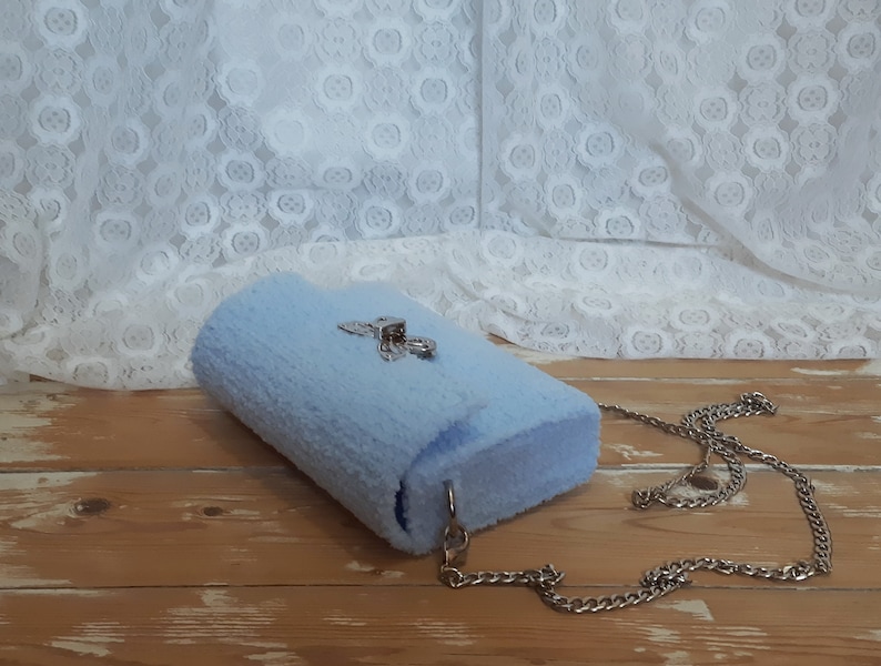 Puede incluir: Un bolso de peluche azul claro con una correa de cadena plateada y un cierre plateado.