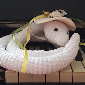 May include: White crocheted snake wearing a straw hat with a yellow ribbon. The snake is sitting on a piano keyboard.