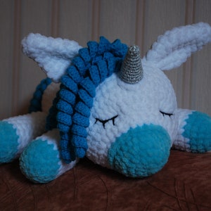 May include: A white crocheted unicorn soft toy with a blue yarn mane and tail, and a silver horn. The unicorn is lying down with its eyes closed.