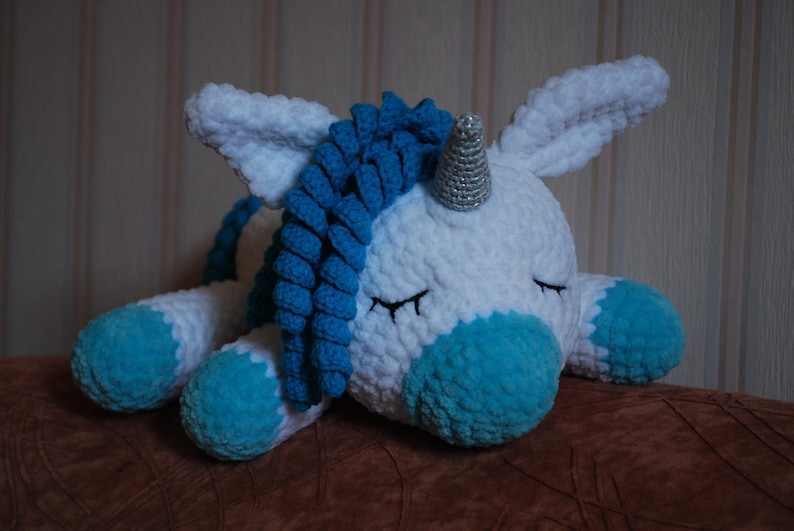 May include: A white crocheted unicorn soft toy with a blue yarn mane and tail, and a silver horn. The unicorn is lying down with its eyes closed.